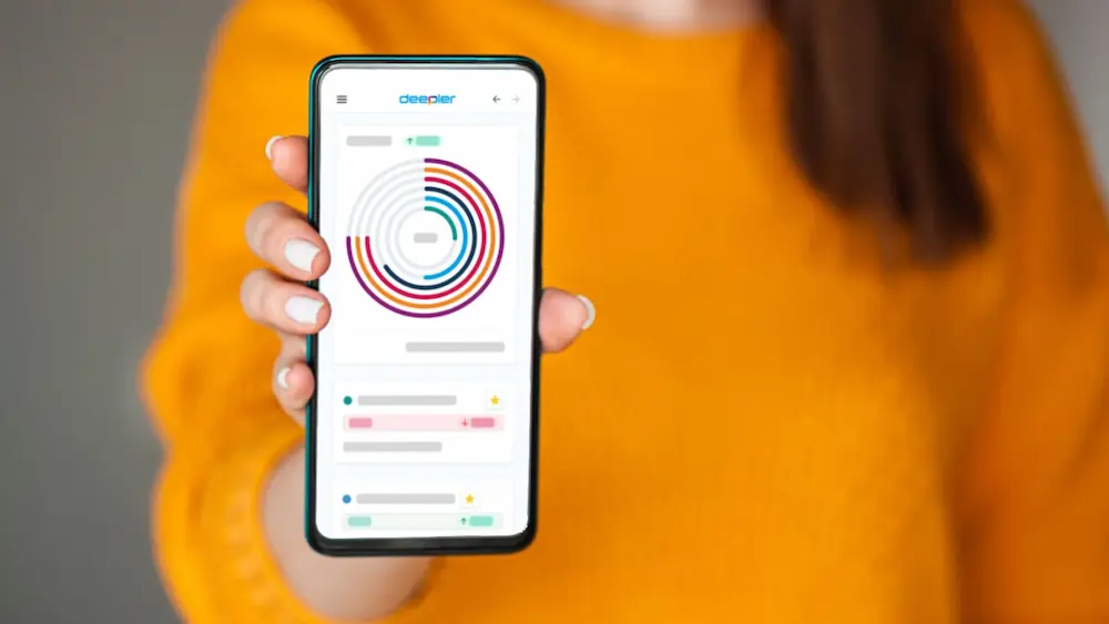 Persoon in gele trui houdt een smartphone vast met de Deeper-app en kleurrijke datavisualisatie op het scherm.