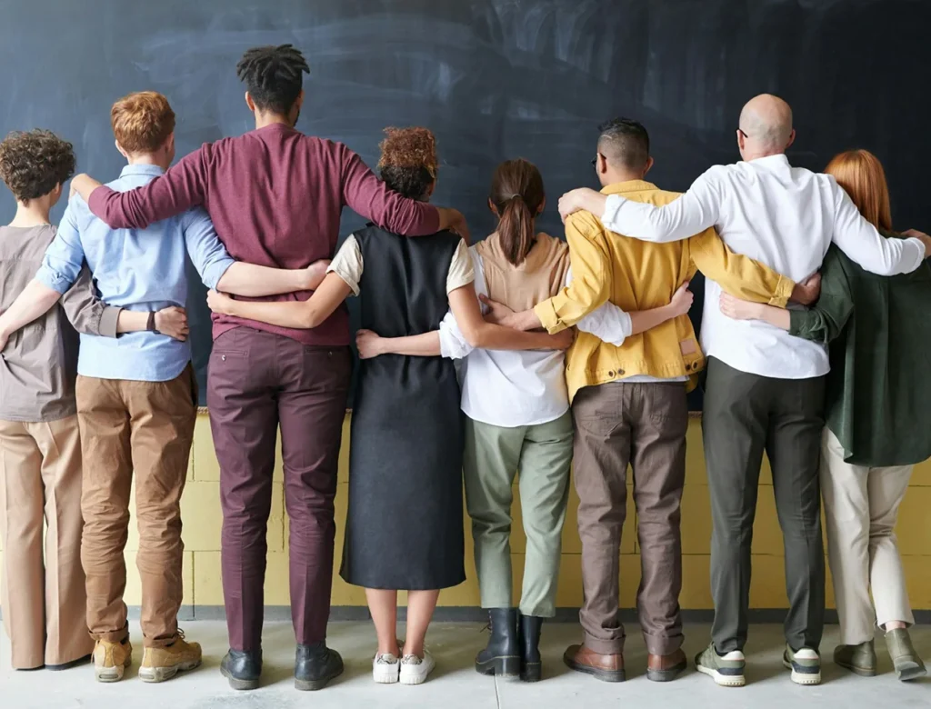 Groep mensen staat zij aan zij met de armen om elkaars schouders, gezien van achteren.