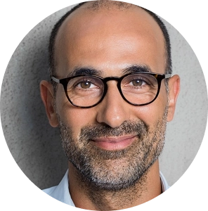 Retrato de un hombre calvo con gafas y barba, sonriendo frente a una pared gris.
