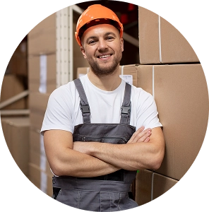 Logistiek medewerker met oranje helm staat glimlachend voor stapels dozen in een magazijn.