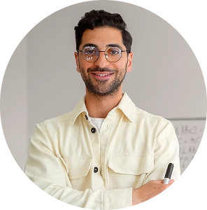 Hombre joven con gafas sonríe mientras sostiene un rotulador en un aula.