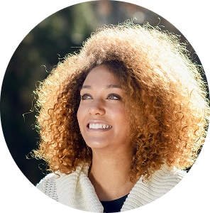Smiling woman with curly hair looking up, wearing a white sweater outdoors.