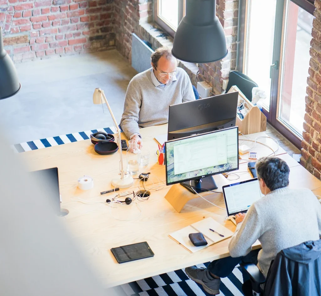 Twee collega’s werken op laptops aan een gedeeld bureau in een modern kantoor met bakstenen muren, van bovenaf gezien.