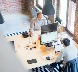 Twee collega’s werken op laptops aan een gedeeld bureau in een modern kantoor met bakstenen muren, van bovenaf gezien.