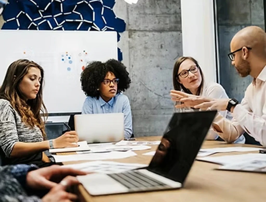 Vier collega’s in een moderne vergaderruimte in gesprek rond een tafel met laptops en documenten.