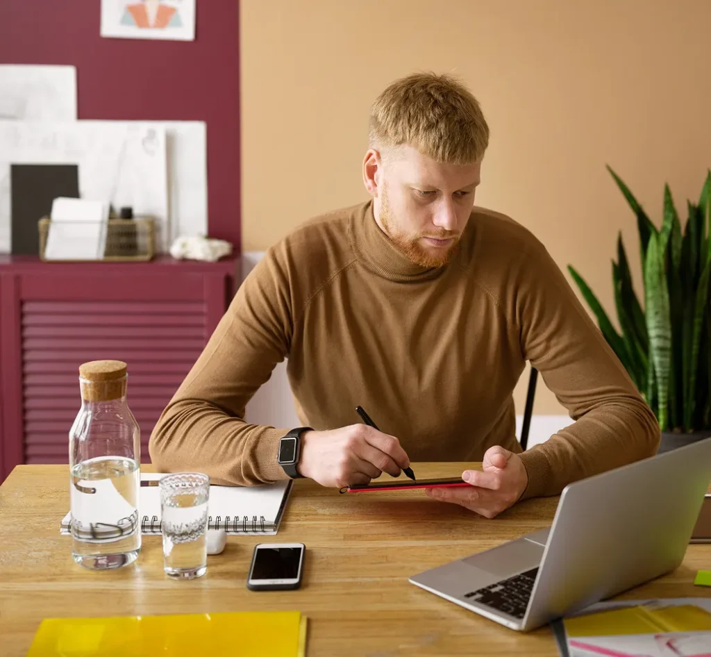 Jonge man werkt geconcentreerd aan een bureau met een tablet, laptop en notitieblok.