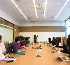Een docent geeft uitleg aan een groep aandachtige studenten in een klaslokaal met laptops rond een vergadertafel.