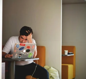 Man werkt geconcentreerd en licht gestrest achter een laptop vol programmeerstickers in een rustige werkruimte.