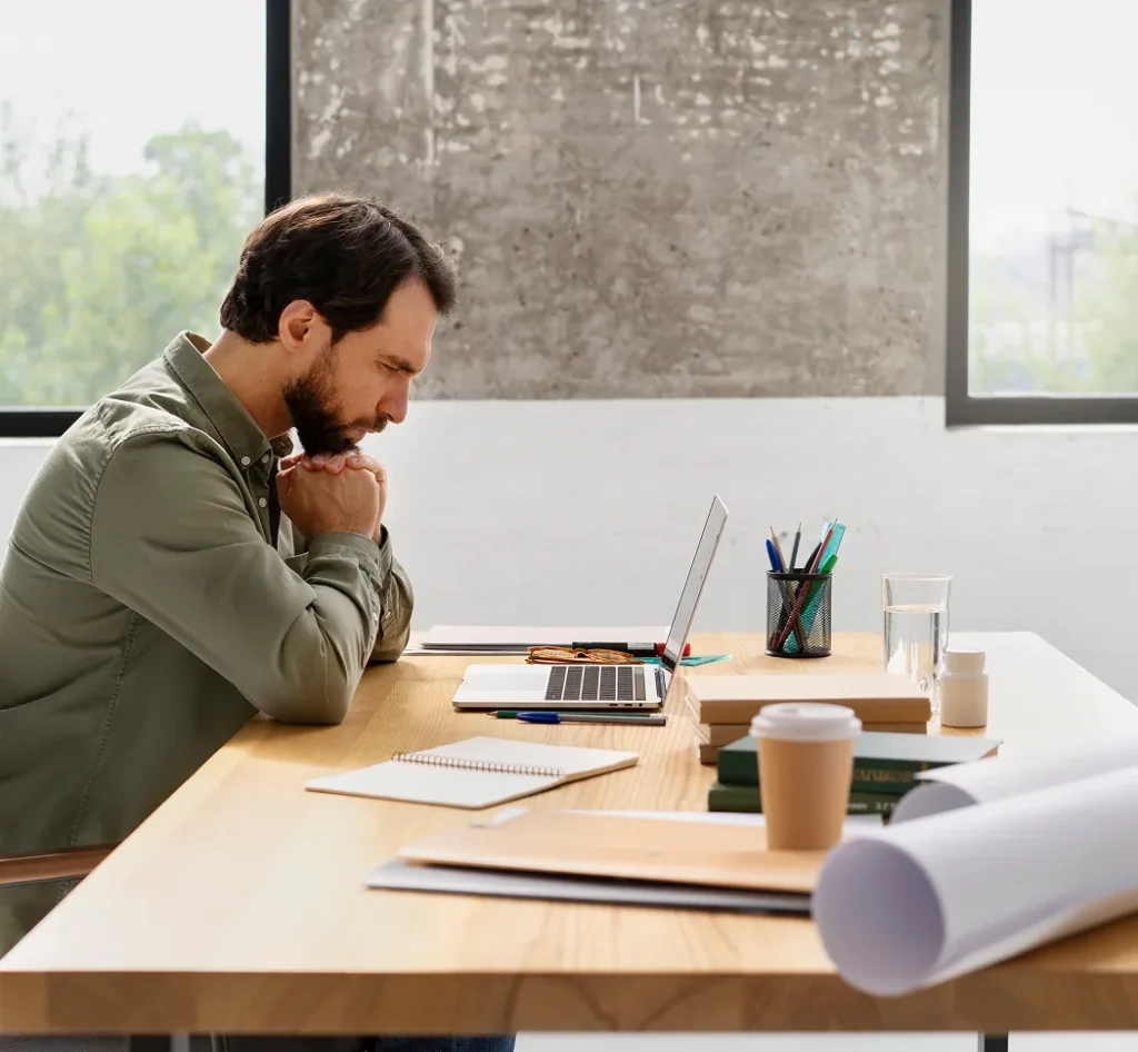 Man leunt voorover aan een bureau terwijl hij geconcentreerd naar een laptop kijkt.