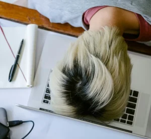 Man met geblondeerd haar ligt met zijn hoofd gefrustreerd op een laptop tussen papieren en een pen.