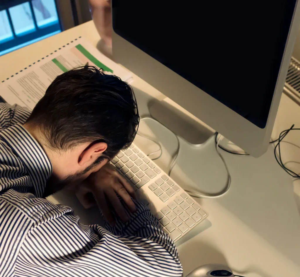 Man met het hoofd op het toetsenbord zittend aan een bureau, ogenschijnlijk teneergeslagen of uitgeput.