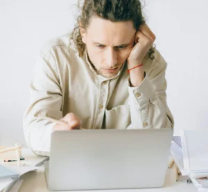 Vermoeide man kijkt geconcentreerd naar zijn laptop aan een rommelig bureau.