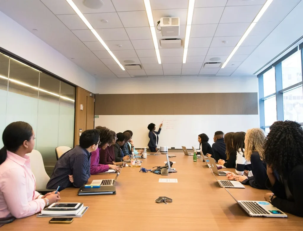 Een groep mensen zit in een vergaderruimte en luistert aandachtig naar een docent die iets uitlegt op een whiteboard.