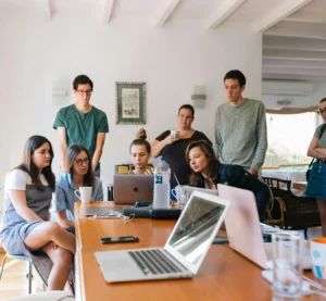 Een team van acht personen kijkt samen aandachtig naar een laptop tijdens een meeting.