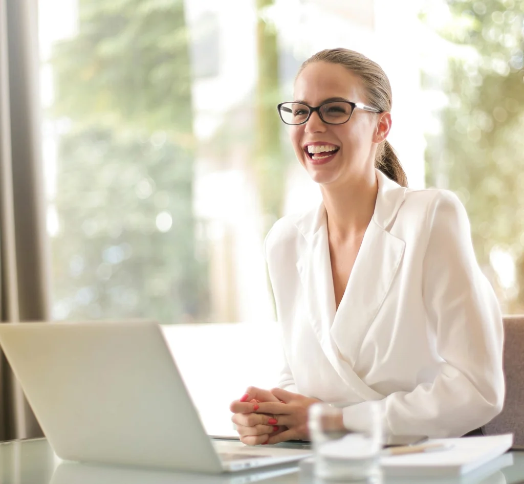 Lachende vrouw met bril achter een laptop aan een bureau in een lichte kantoorruimte.