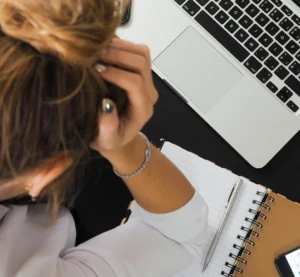 Vrouw met hoofd in haar hand zit teneergeslagen aan bureau met laptop en notitieboek.