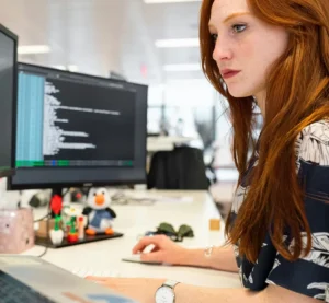 Vrouw werkt geconcentreerd achter twee computerschermen met programmeercode op een modern kantoor.