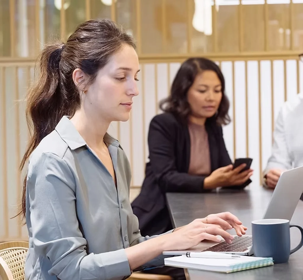 Vrouw in grijze blazer werkt op laptop aan vergadertafel met collega's op de achtergrond.